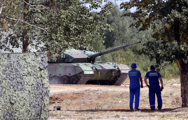 Ein getarnter Panzer steht im Gelände, beobachtet von zwei Personen in blauen Overalls.