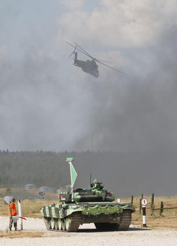 Ein grüner Panzer steht auf einem Feld, während ein Hubschrauber in der Ferne fliegt.