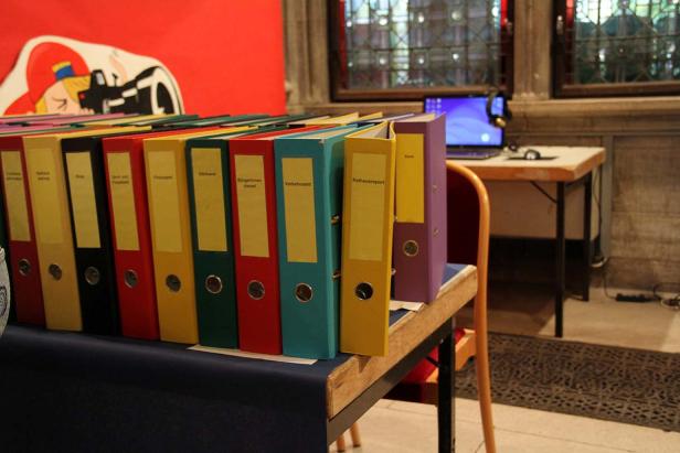 Ein Tisch mit farbigen Aktenordnern und Beschriftungen wie „Finanzamt“ und „Rathausreport“.