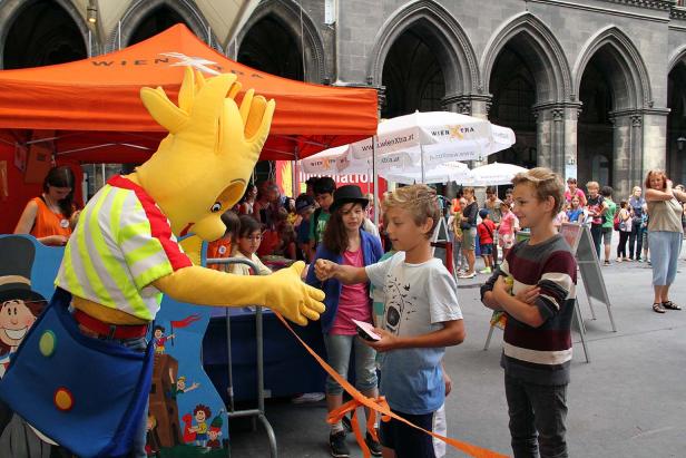Ein gelbes Maskottchen gibt einem Jungen die Hand vor einem Stand von WienXtra.