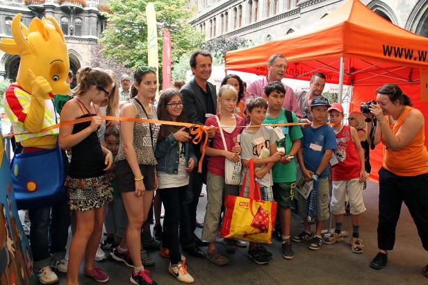 Eine Gruppe von Kindern und Erwachsenen steht vor einem orangefarbenen Zelt und hält ein Band fest.
