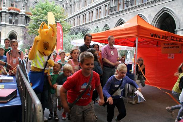 Eine Gruppe Kinder läuft an einem Stand mit einem Maskottchen vorbei.