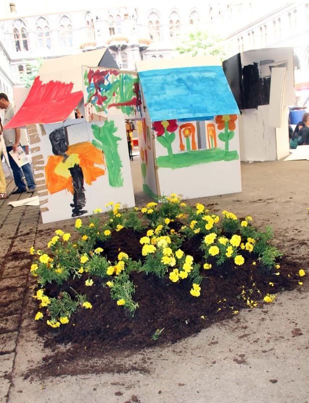 Gelbe Blumen vor bemalten Papphäusern auf einem Platz.