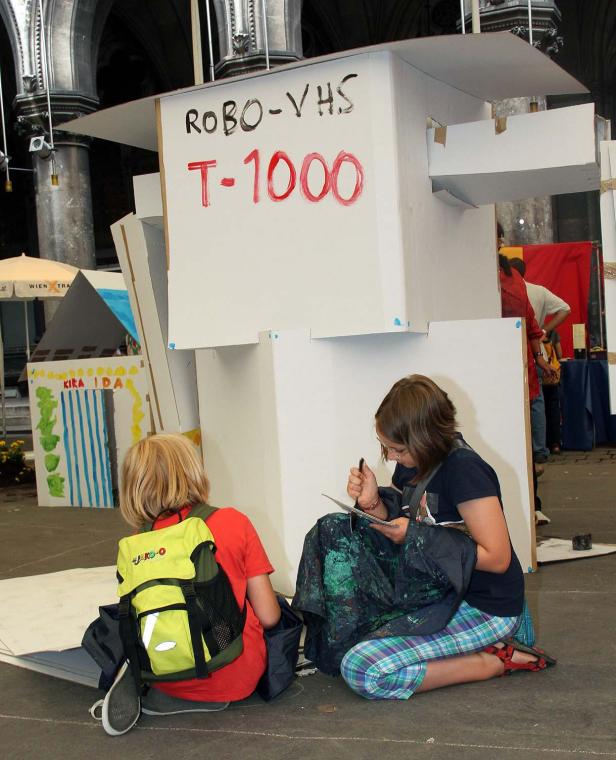 Zwei Kinder sitzen vor einem Papproboter mit der Aufschrift „Robo-VHS T-1000“.