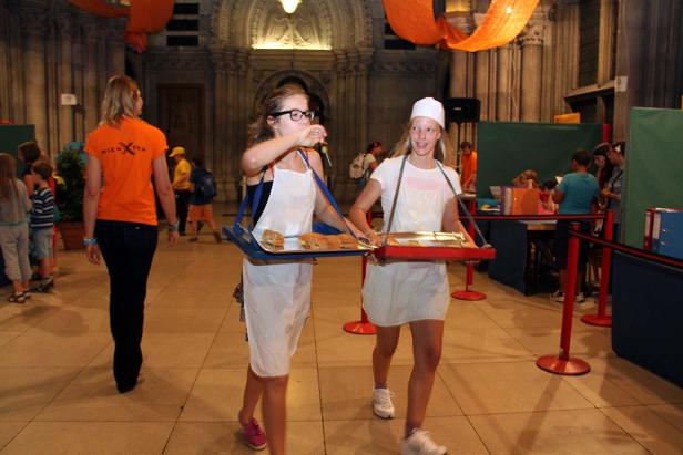 Zwei Mädchen in Kochschürzen tragen Tabletts mit Essen in einem belebten Raum.