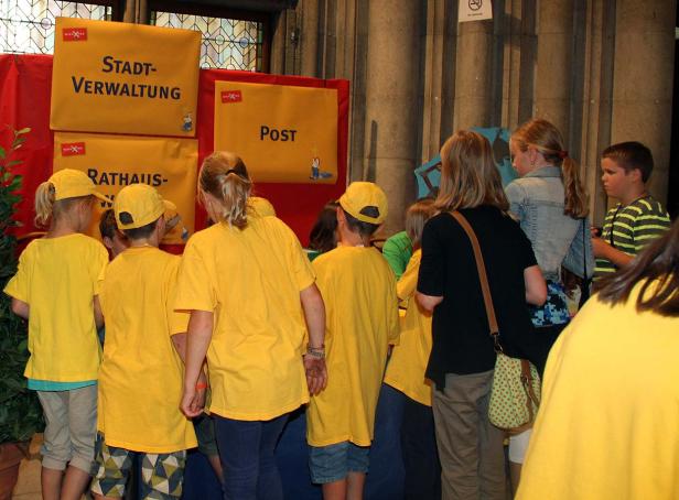 Eine Gruppe Kinder in gelben T-Shirts steht vor Schildern mit Aufschriften wie „Stadtverwaltung“ und „Post“.