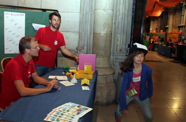 Ein Mädchen mit Hut steht vor einem Stand mit zwei Männern in roten T-Shirts.