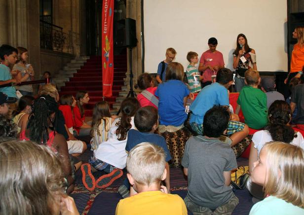 Eine Gruppe von Kindern sitzt vor einer Leinwand, während zwei Personen sprechen.