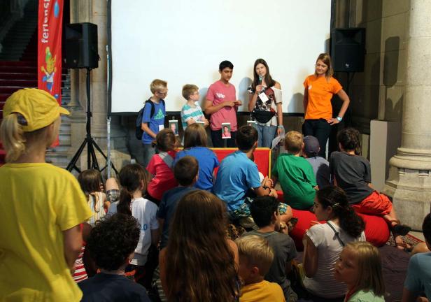 Eine Gruppe Kinder sitzt vor einer Bühne, auf der drei Personen stehen und sprechen.