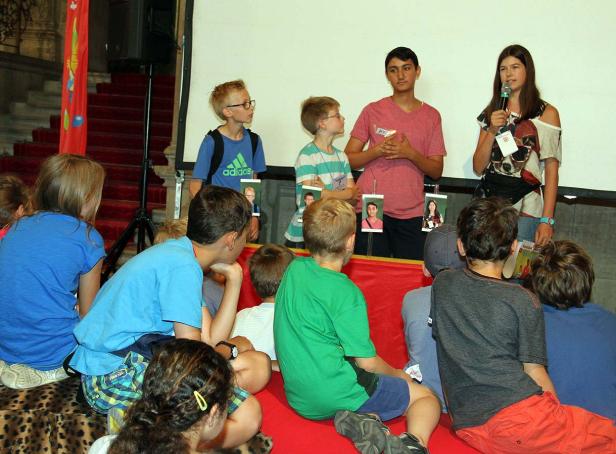 Eine Gruppe Kinder sitzt vor einer Leinwand, während zwei Jugendliche sprechen.