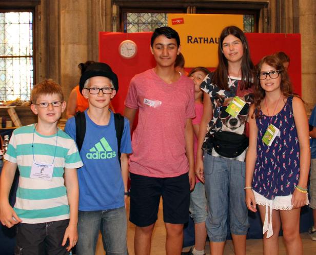 Eine Gruppe von Kindern steht vor einem roten Hintergrund mit der Aufschrift „Rathaus“.