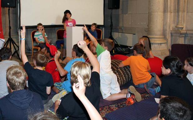 Eine Gruppe Kinder sitzt und hört aufmerksam zwei Vortragenden zu.