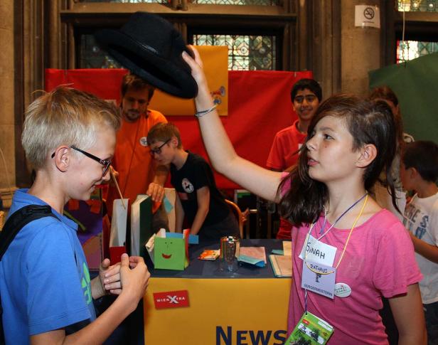 Ein Mädchen hält einen Hut hoch, während ein Junge zusieht; im Hintergrund weitere Kinder und Erwachsene.