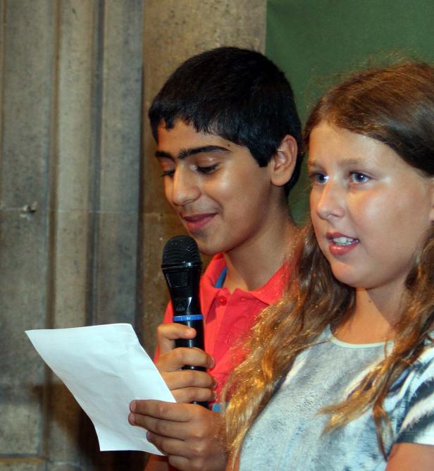 Ein Junge liest mit einem Mikrofon in der Hand von einem Blatt Papier, neben ihm ein Mädchen.