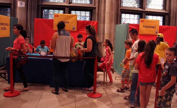 Kinder stehen Schlange vor Ständen mit Aufschriften wie „Bank“ und „Finanzamt“.