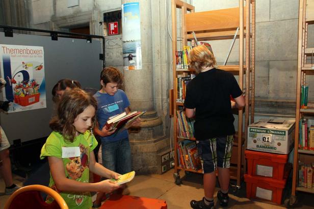 Kinder suchen Bücher in einer Bibliothek aus Regalen auf Rollen.