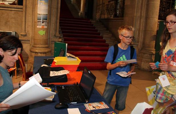 Eine Frau liest Dokumente an einem Tisch, während zwei Jugendliche sich unterhalten; im Hintergrund eine Treppe mit rotem Teppich.