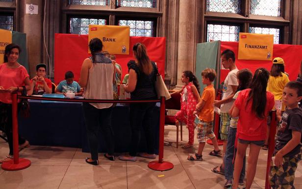Kinder stehen an einem Stand mit den Aufschriften „Bank“ und „Finanzamt“.
