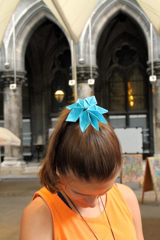 Eine Frau trägt eine blaue Origami-Blume im Haar vor einem Gebäude mit Torbögen.