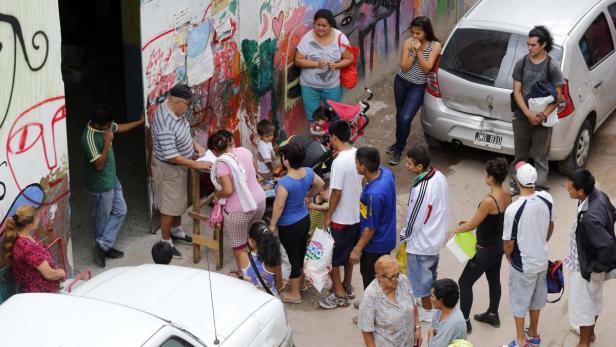 Auch wenn der Andrang bei Suppenküchen wie dieser in Buenos Aires groß ist: Im Vergleich zu 2002 ist es diesmal auf den Straßen ruhig. Eine Gruppe von Menschen steht vor einem Gebäude mit Graffiti an der Wand.