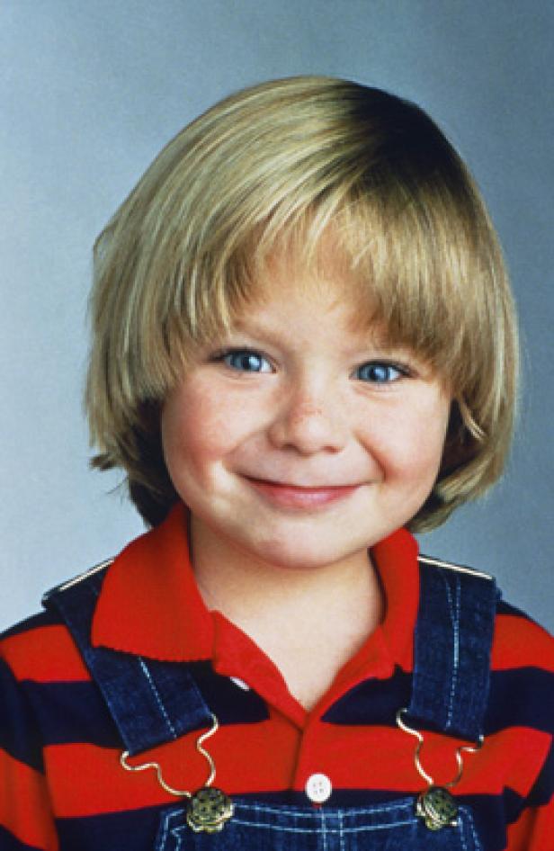 Ein lächelnder Junge mit blonden Haaren und blauen Augen trägt eine gestreifte Latzhose.