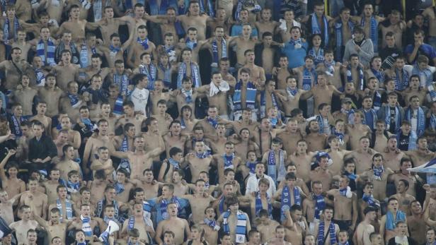 Eine große Gruppe Fußballfans jubelt mit nacktem Oberkörper und Schals im Stadion.