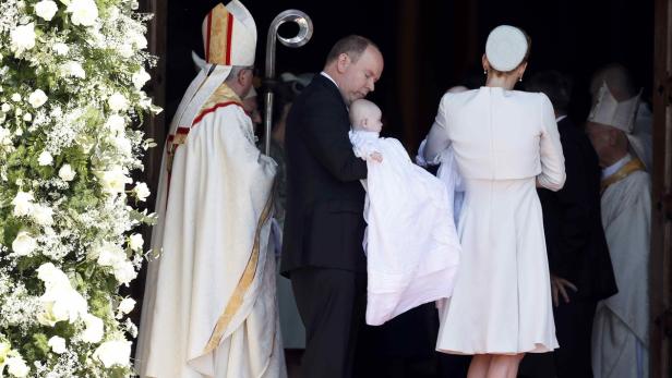 Fürst Albert von Monaco hält ein Baby in den Armen, während eine Frau in Weiß daneben steht.