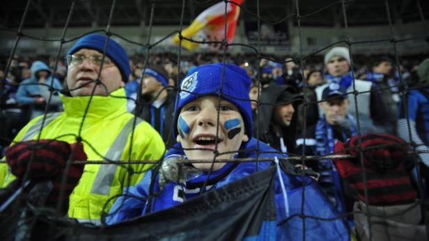 Ein junger Fan mit blau-schwarzer Gesichtsbemalung schaut durch einen Zaun bei einem Fußballspiel.