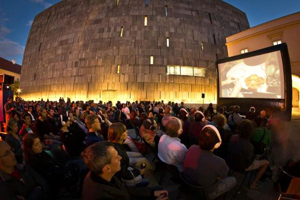 Eine Open-Air-Kinovorstellung mit vielen Zuschauern vor einem großen Gebäude.