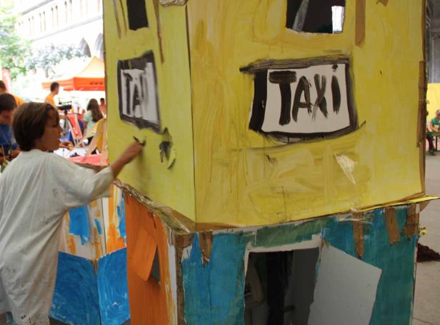 Ein Kind bemalt ein gelbes Papphaus mit der Aufschrift „Taxi“.