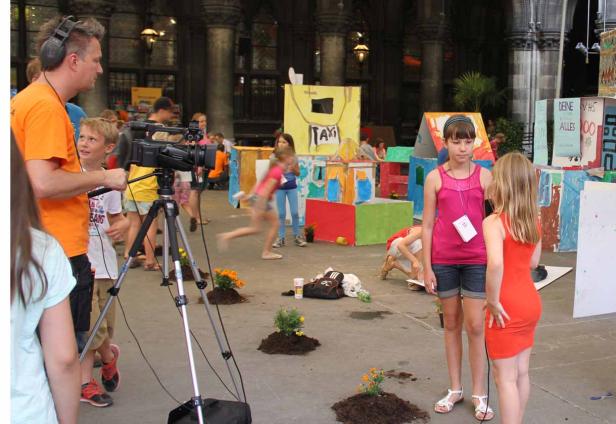 Ein Kameramann filmt Kinder vor einer Kulisse aus Papphäusern.