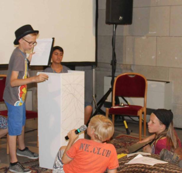 Eine Gruppe von Kindern führt ein Theaterstück mit Mikrofon und Rednerpult auf.
