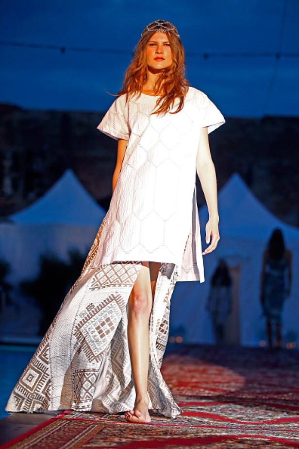 Eine barfüßige Frau in einem weißen Kleid mit geometrischen Mustern läuft über einen roten Teppich.