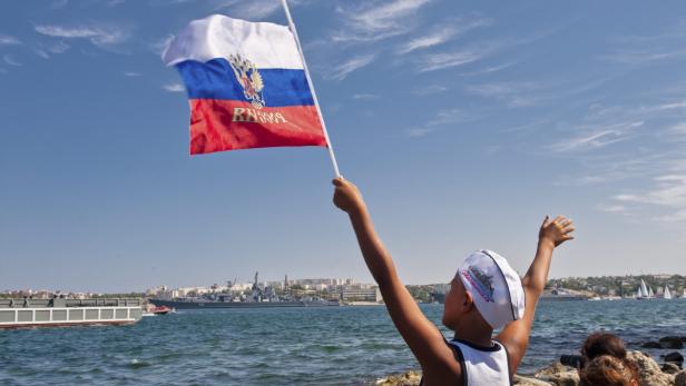 Ein Junge schwenkt eine russische Flagge am Ufer, im Hintergrund sind Schiffe zu sehen.