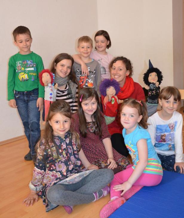 Eine Gruppe von Kindern und eine Frau sitzen zusammen mit Puppen in einem Raum.