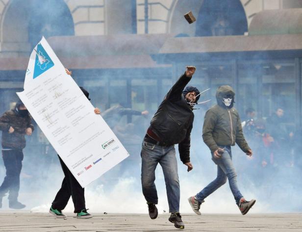 Bei einer Demonstration wirft ein Mann einen Stein, während andere Rauchbomben zünden und ein großes Schild tragen.