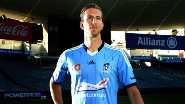 Ein Mann im hellblauen Trikot von Sydney FC posiert in einem Stadion.
