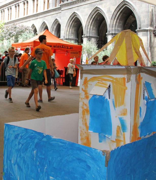 Kinder bemalen einen Pappkarton vor einem Gebäude in Wien.
