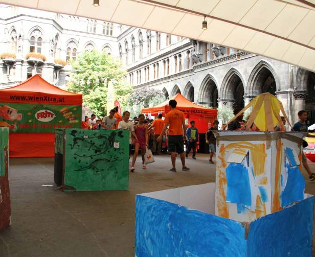 Kinder spielen auf einem Platz mit bemalten Kartons und Zelten im Hintergrund.
