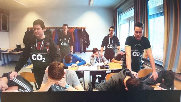 Schüler mit CO2-Symbolen auf ihren T-Shirts stehen um eine Gruppe erschöpfter Schüler herum.