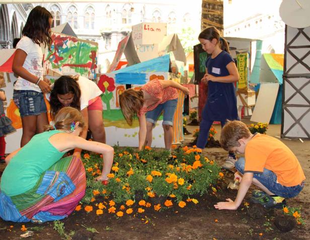 Eine Gruppe Kinder pflanzt orangefarbene Blumen vor einer Kulisse aus bemalten Papphäusern.
