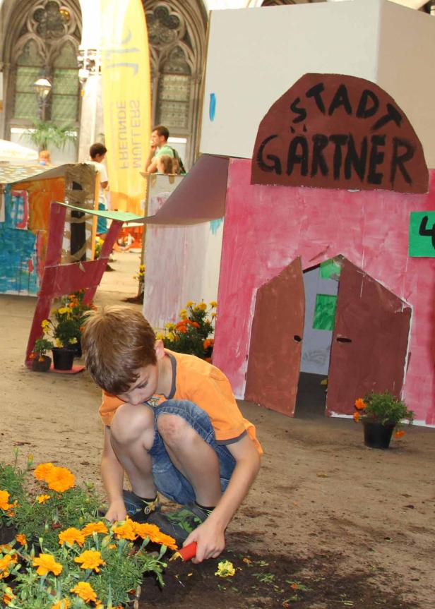 Ein Junge bepflanzt ein Beet mit Ringelblumen vor einem bemalten Papphaus mit der Aufschrift „Stadtgärtner“.