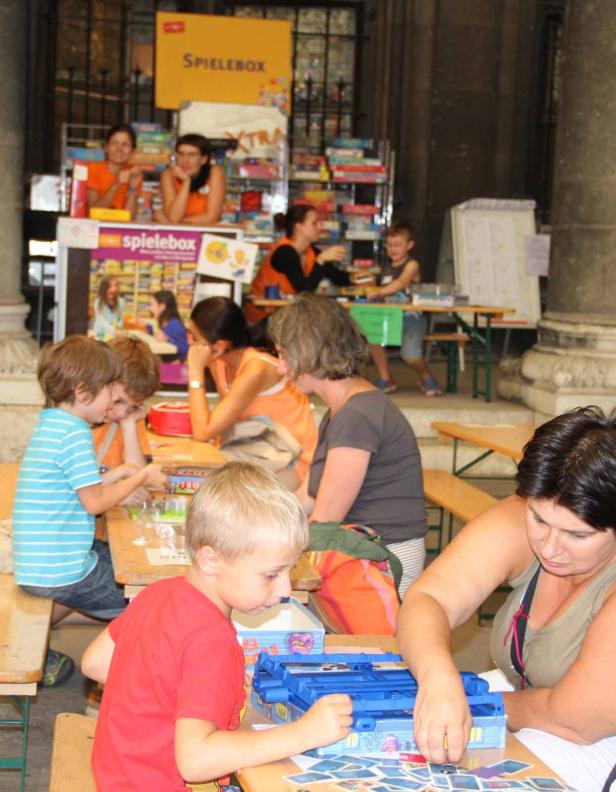 Kinder und Erwachsene spielen zusammen an Tischen bei einer „Spielebox“-Veranstaltung.