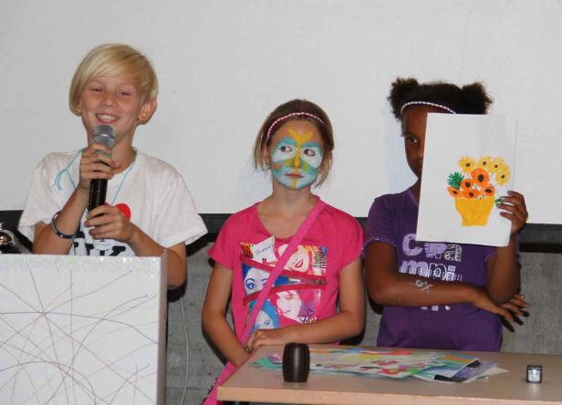 Drei Kinder stehen an einem Tisch, eines mit Gesichtsbemalung und eines mit einer Zeichnung.