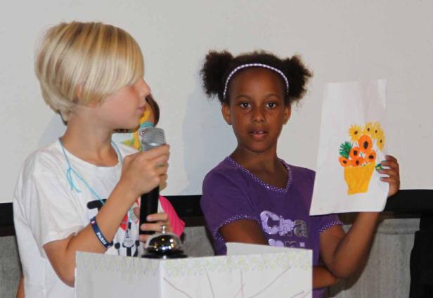 Zwei Kinder stehen an einem Tisch, eines hält ein Mikrofon, das andere eine Zeichnung von Blumen.