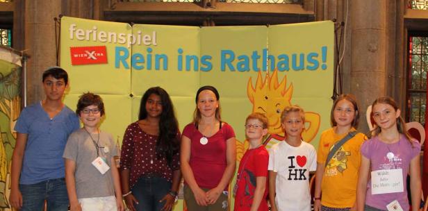 Eine Gruppe von Kindern steht vor einem Banner für das „Ferienspiel Rein ins Rathaus!“.