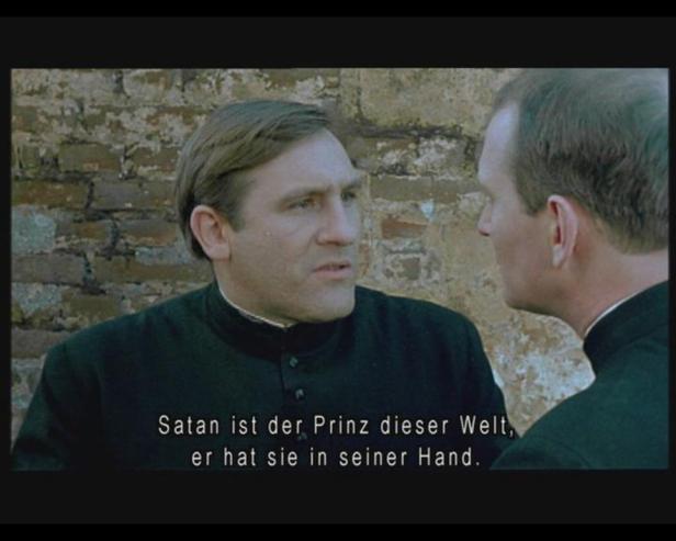 Zwei Männer in Priesterkleidung unterhalten sich vor einer Ziegelmauer. Im Vordergrund steht der Text: „Satan ist der Prinz dieser Welt, er hat sie in seiner Hand.“.