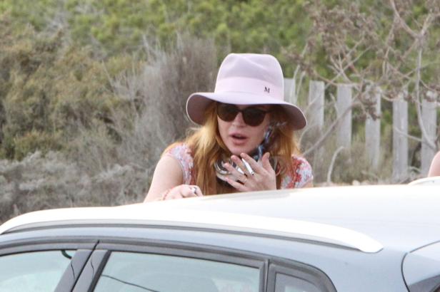Lindsay Lohan mit Hut und Sonnenbrille raucht eine Zigarette am Auto.