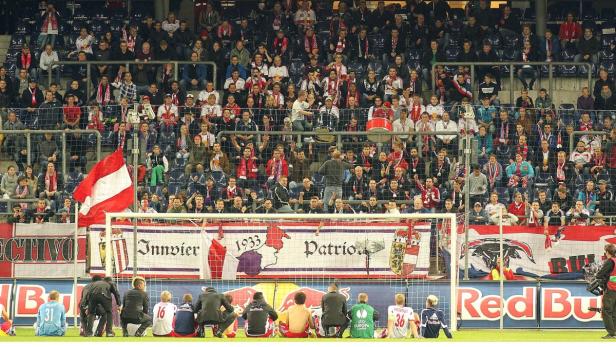 Fußballfans jubeln im Stadion hinter dem Tor, einige Spieler sitzen auf dem Rasen.