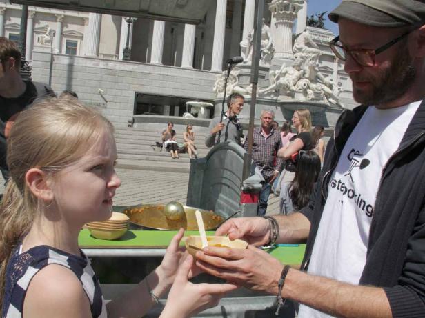 Ein Mann reicht einem blonden Mädchen eine Schale mit Essen vor dem Parlament in Wien.